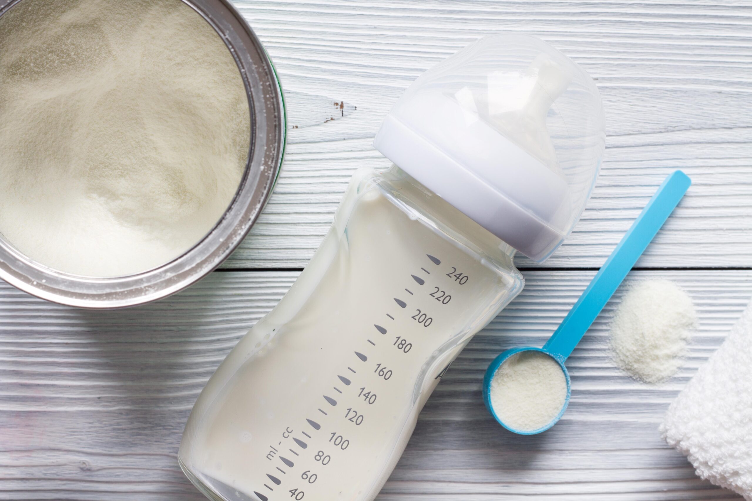 Baby bottle with prepared infant formula beside a scoop and container of powdered formula on a wooden surface.